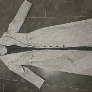 Grå lång kappa med knappar - Snygg grå kappa med klassisk krage och svarta knappar framtill. Lång modell med två fickor och slits bak. Fodrad insida i svart tyg. Perfekt för dig som gillar stilrena och tidlösa plagg.