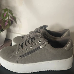 Grå sneakers med dragkedja Everest - Snygga grå sneakers från Everest med plattformssula och rund tå. Skorna har snörning och en cool dragkedjedetalj på sidan samt metallic-silver på hälen. Materialet är mockaliknande syntet och insidan är mjuk för extra komfort. Perfekt för dig som gillar stilrena och trendiga skor.