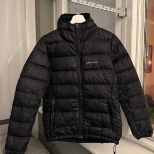 Svart dunjacka Peak Performance - Svart dunjacka från Peak Performance med huva och dragkedja framtill. Jackan har inga defekter mer än hämpan (se bild 5), två sidofickor med dragkedja och en innerficka. Perfekt för kalla dagar och har en clean, sportig look.
