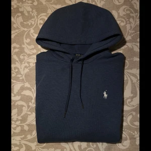 Marinblå hoodie från Polo Ralph Lauren - Snygg marinblå hoodie från Polo Ralph Lauren med klassisk vit broderad logga på bröstet. Tröjan har huva med snörning, stor magficka och långa ärmar. Mjuk bomullskvalitet som är skön att ha på sig. Perfekt för dig som gillar stilrena och bekväma plagg. ❗️M passar S❗️