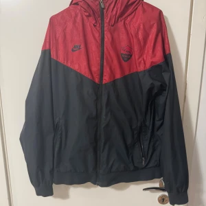 Nike AS Roma röd-svart vindjacka - Snygg vindjacka från Nike i AS Roma-design med röd övre del och svart nedre del. Jackan har huva, dragkedja framtill och två sidofickor med dragkedja. Röd del med subtilt mönster och AS Roma-logga på bröstet. Perfekt för dig som gillar fotboll och streetwear. Limited editon Roma x Nike