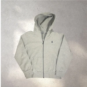 Grå hoodie från Polo Ralph Lauren - Snygg ljusgrå hoodie från Polo Ralph Lauren med dragkedja framtill och klassisk broderad logga på bröstet. Mycket bra skick, inga hål. Man kan även addera snören till tröjan om man vill✅ skickar snabbt efter att du har beställt📦