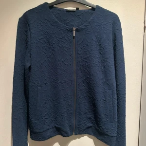 Mörkblå cardigan jacka från KappAhl - Snygg mörkblå cardigan jacka från KappAhl med strukturmönster i tyget. Jackan har rund halsringning, lång ärm och dragkedja framtill. 