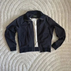 Mörkblå bomberjacka Peak Performance - Snygg mörkblå bomberjacka från Peak Performance med klassisk krage och ribbade muddar. Jackan har dragkedja framtill och är tillverkad i bomull och polyamid, med ljus insida i nylon. Perfekt för dig som gillar stilren och sportig look.