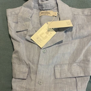 Ljusblå linneskjorta från Dressmann - Fräsch ljusblå skjorta från Dressmann i premium linne. Skjortan har korta ärmar, klassisk krage, två bröstfickor med lock och knäppning framtill. Perfekt för varma dagar och har en chill, stilren vibe.