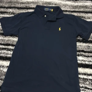 Marinblå pikétröja från Ralph Lauren - Snygg marinblå pikétröja från Polo Ralph Lauren med klassisk krage och två knappar. Ikoniska gula loggan broderad på bröstet. Tröjan är i bomull och har korta ärmar, perfekt för en clean och stilren look.