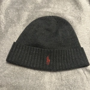 Mörkgrå merinoullsmössa Polo Ralph Lauren - Snygg mörkgrå mössa från Polo Ralph Lauren i mjuk och tvättbar merinoull. Klassisk ribbad kant och röd broderad Polo-logga framtill. Perfekt för kalla dagar och enkel att matcha med din stil.