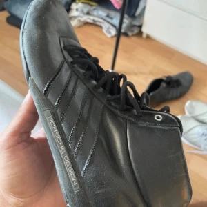 Adidas Porsche Design svarta sneakers - Svarta Adidas Porsche Design sneakers med högt skaft och snygga detaljer. Skorna har snörning, rund tå och är tillverkade i skinn med diskret Adidas-logga på sidan. Stilren och sportig look som passar dig som gillar exklusiva samarbeten. Limited edition skor