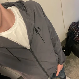 Grå vindjacka från Under Armour - Snygg grå vindjacka från Under Armour med huva och svarta snören. Jackan har dragkedja framtill, två sidofickor och en stilren logga på bröstet. Perfekt för dig som gillar sportig och clean stil. Passar till jeans eller träningskläder. Pris kan diskuteras vid snabbaffär