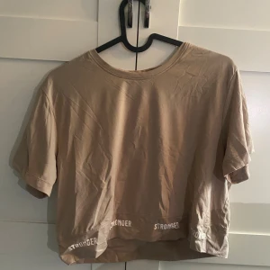 Beige croppad topp från Stronger - Säljer en beige croppad topp från Stronger med korta ärmar och rund hals. Nedtill finns vit text med loggan 'STRONGER' runt om. Materialet är mjukt och stretchigt, skönt att träna i. Finns matchande byxor!