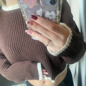 Brun croppad stickad tröja - Mysig brun stickad tröja med croppad passform och vita ribbade kanter vid hals, ärmslut och nederkant. Perfekt för lager-på-lager och chill dagar. Tröjan har lång ärm och är ribbstickad för extra struktur.