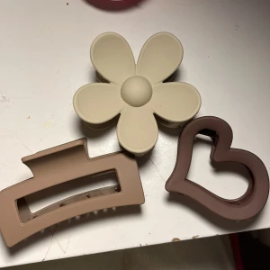 Tre hårklämmor i beige och brunt - Säljer tre hårklämmor i olika former: en beige blomma, en ljusbrun rektangulär och en brun hjärtformad klämma. Perfekta för att sätta upp håret med en söt och trendig touch. Passar dig som gillar stilrena accessoarer i neutrala färger.