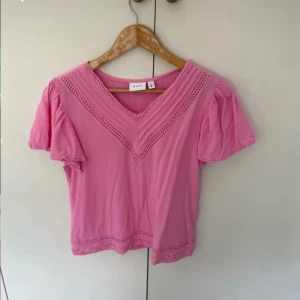 Rosa blus med spets från Vila - Säljer en söt rosa blus från Vila med v-ringad hals och dekorativa spetsdetaljer längs halsringning och nederkant. Blusen har korta puffärmar och är gjord i mjuk bomull. Perfekt för dig som gillar färg och romantiska detaljer!