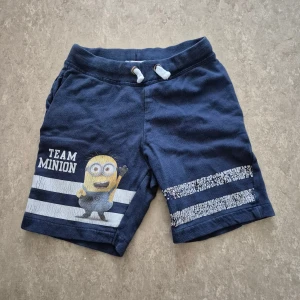 Shorts - Snygga mörkblå shorts från H&M med Minion-tryck och texten 'TEAM MINION' på ena benet. De har vita snören i midjan och coola vita ränder med slitna detaljer. Perfekt för dig som gillar bekväma och lekfulla plagg.