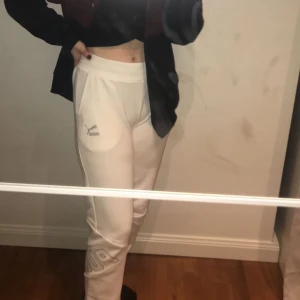 Vita mjukisbyxor Puma - Snygga vita mjukisbyxor från Puma med logga på låret och tryck på benet. Byxorna har en normal passform och är tillverkade i mjukt material som är perfekt för chill eller träning. Elastisk midja och bekväma fickor på sidorna.