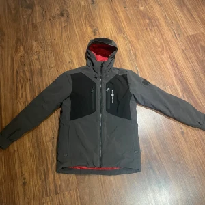 Grå och svart vinterjacka från sail racing - Säljer en grå och svart vindjacka från sail racing med huva och rödfärgat innerfoder. Jackan har dragkedja framtill, två bröstfickor med dragkedja och stilrena paneler i svart på bröst och rygg. Perfekt för dig som gillar funktionell och snygg design.