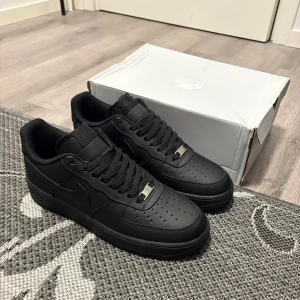 Nike Air Force 1 helsvarta sneakers - Nike Air Force 1 sneakers i helsvart färg med klassisk låg siluett. Skorna har en platt sula, rund tå och är tillverkade i skinn med perforerade detaljer på tån för extra ventilation. Snygga snörningar och ikonisk Nike-logga på sidan. Perfekta för dig som gillar stilrena och tidlösa sneakers.