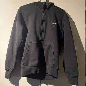 Svart hoodie Comme des Garçons Play - Svart hoodie från Comme des Garçons Play med halv dragkedja och ikoniskt hjärta med ögon på bröstet och stort tryck på ryggen. Mjuk bomull, känguruficka och ribbade muddar. Perfekt för dig som gillar streetwear och stilrena detaljer. Storlek S men passar M. Använd endast 2 gånger.