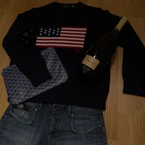 Mörkblå stickad tröja Polo Ralph Lauren - Mörkblå stickad tröja från Polo Ralph Lauren med amerikansk flagga och RL-broderi på bröstet. Klassisk rund halsringning och ribbade muddar. Perfekt för dig som vill ha en stilren och ikonisk look.