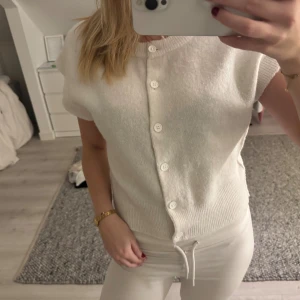 Vit stickad kortärmad blus med knappar - Säljer en söt vit stickad blus med korta ärmar och rund hals. Blusen har knäppning framtill med vita knappar och ribbad nederkant. Perfekt för en clean och stilren look.