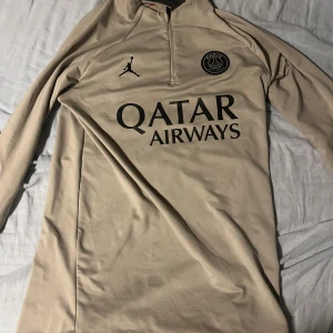 Beige PSG långärmad fotbollströja - Säljer en beige långärmad PSG fotbollströja från Jordan med svart Paris Saint-Germain-logga och Qatar Airways-tryck på bröstet. Tröjan har halv dragkedja och snyggt Paris-tryck på ärmen. Materialet är mjukt och stretchigt, perfekt för träning eller match.