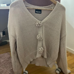 Beige stickad kofta från Pieces - Supermysig beige stickad kofta från Pieces med v-ringad hals och tre stora, virkade blomknappar framtill. Koftan har långärmad passform och ribbade muddar vid ärmslut och nederkant. Perfekt för lager-på-lager och chill dagar. Använd  3 gånger kanske 