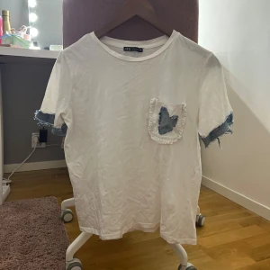 Vit t-shirt med denimdetaljer från Zara - Säljer en vit t-shirt från Zara med coola denimdetaljer på ärmarna och en ficka framtill med fransad kant och blå denimpatch. T-shirten har rund hals och avslappnad passform, perfekt för en chill och trendig look. ! denna tshirt är omsydd och inte köpt så!