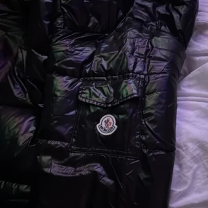 Svart Moncler pufferjacka med huva - Säljer en svart pufferjacka från Moncler i storlek L. Jackan har en glansig finish, huva, dragkedja framtill och ficka med Moncler-logga på ärmen. Perfekt för kalla dagar och har en riktigt snygg streetstil. Oanvänd jacka!