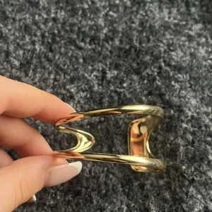 Guldigt öppet armband i metall - Snyggt och modernt armband i guldtonad metall med en öppen, böjd design. Armbandet har två parallella band som ger en cool och unik look. Perfekt för dig som gillar minimalistiska och stilrena accessoarer.