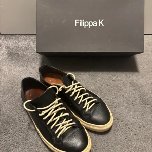  Filippa K sneaker - Snygga svarta sneakers från Filippa K med rund tå och platt beige sula. Skorna är tillverkade i skinn och har vita snören som kontrast mot det svarta lädret. Klassisk och stilren design som passar till många olika looks.