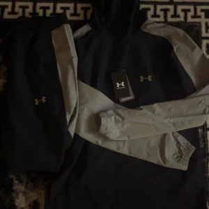 Under armour tracksuite helt ny  passar storlek S/M - Snygg vindjacka från Under Armour i svart och grått med reflekterande loggor. Jackan har huva, dragkedja och elastiska muddar vid ärmslut. Perfekt för träning utomhus och har en sportig vibe. Materialet är lätt och syntetiskt för maximal komfort.