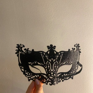 Svart maskeradmask med spetsdetaljer - Säljer en svart maskeradmask med snygga spetsiga detaljer och elegant mönster. Masken har ett elastiskt band för att sitta bekvämt runt huvudet. Perfekt för maskerad eller temafest när du vill sticka ut med en mystisk look.