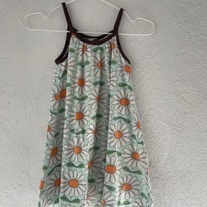 Blommig sommarklänning från Lindex - Söt kortklänning från Lindex Kids med blommigt mönster i vitt, orange och grönt. Klänningen har bruna axelband och kant, är ärmlös och gjord i mjuk bomull. Perfekt för soliga dagar och har en luftig passform som gör den skön att bära.