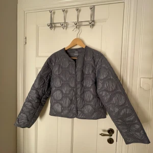 Grå quiltad jacka från MANGO - Quiltad jacka från MANGO i ljusare grå färg med vågigt mönster. Jackan har rund hals, lång ärm. Materialet är syntet och ger en lätt och modern känsla. Köptes för 699 kr. Önskar du bilder med jackan på är det bara att höra av sig så fixar vi det!