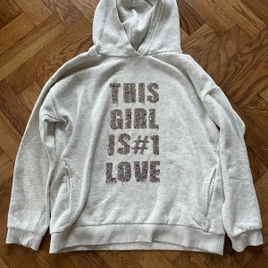 Grå hoodie Zara med paljetttext - Ljusgrå hoodie från Zara Girls i storlek 164. Framsidan har texten 'THIS GIRL IS #1 LOVE' i paljetter som skiftar mellan silver och rosa beroende på hur du drar över dem. Tröjan har huva, långa ärmar och mjukt material i bomullsblandning. Perfekt för dig som gillar statement-plagg!