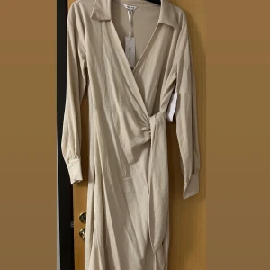 Beige omlottklänning från Happy Holly - Snygg beige långklänning från Happy Holly med omlott-design och knyt i midjan. Klänningen har långärmade ärmar med manschett och knapp, klassisk krage och mjukt bomullsmaterial. Perfekt för dig som gillar stilrena och enkla plagg med en twist.