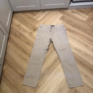 Beige chinos från Dressmann slim fit - Snygga beige chinos från Dressmann i slim fit-modell. Klassisk design med framfickor, bakfickor med knapp och bälteshällor. Tillverkade i mjukt bomullsmaterial som är skönt mot huden. Perfekt för dig som gillar stilrena och enkla byxor.