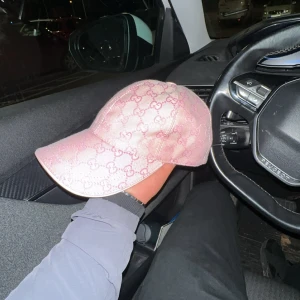 Rosa Gucci keps med GG-mönster - Helt oanvänd och snygg rosa keps från Gucci med ikoniskt GG-mönster över hela kepsen. Justerbar rem baktill och böjd skärm. Tillverkad i slitstark bomullsmix med broderade detaljer. Perfekt statement-piece för dig som gillar lyxiga accessoarer.