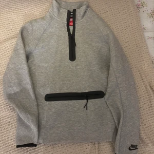 Grå half zip tröja från Nike - Snygg grå tröja från Nike med svart logga på ärmen. Tröjan har half zip framtill, hög krage och en stor ficka med dragkedja över magen. Perfekt för träning eller chill. Tillverkad i mjukt material med svarta detaljer.