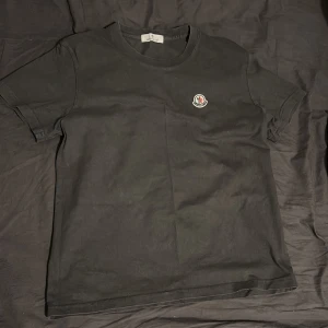 Svart Moncler t-shirt med logga - Svart t-shirt från Moncler i mjuk bomull med klassisk rund hals och korta ärmar. På bröstet sitter Monclers ikoniska broderade logga i vitt, blått och rött. Enkel och stilren design som funkar till allt.
