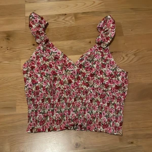 Blommig croppad topp - Supersöt croppad linnetopp med blommigt mönster. Toppen har volangdetaljer på axelbanden och smockad midja och öppen rygg. I nyskick. 