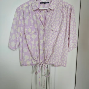 Ljuslila blommig skjorta French Connection - Säljer en ljuslila skjorta från French Connection med blommigt mönster i vitt och gult. Skjortan har knappar framtill, två bröstfickor och knytning i midjan. Ärmarna är trekvarts och modellen är lite croppad för en chill vibe.