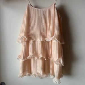 Champagnefärgad plisserad kortklänning - Säljer en champagnefärgad kortklänning med tunna axelband och plisserat tyg. Klänningen har volangdetaljer i flera lager och ett skirt, luftigt material som ger en romantisk vibe. Perfekt för dig som gillar en feminin och trendig look.