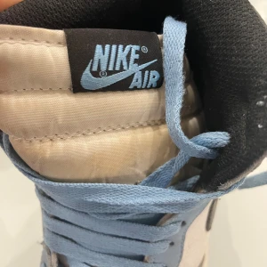 Nike Air Jordan 1 High blå/vit - Nike Air Jordan 1 High sneakers med ljusblå mockadetaljer, vit skinnpanel och svarta swoosh-loggor. Klassisk high-top siluett med blå snörning och rund tå. Yttersulan är blå och mellansulan vit. Perfekt för dig som gillar streetstyle och ikoniska sneakers.