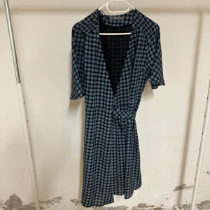 Blommig omlottklänning från Fiore - Snygg omlottklänning från Fiore med svart botten och blått  mönster. Klänningen har v-ringning, knyt i midjan och korta ärmar. Längden är något längre än knälång och materialet känns lätt och luftigt, perfekt för dig som gillar stilrena detaljer.