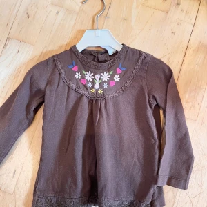 Brun blus med broderade blommor Zara - Supersöt brun blus från Zara med broderade blommor och hjärtan i färgglada nyanser vid halsen. Blusen har långärmad design och fina detaljer längs kragen och nederkanten. Perfekt för dig som gillar gulliga och lekfulla plagg till de små.