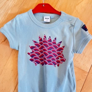 Ljusblå t-shirt från P.O.P med tryck - Säljer en ljusblå t-shirt från P.O.P i mjuk bomull. T-shirten har korta ärmar och ett coolt grafiskt tryck i rött, lila och vitt på bröstet. Rund hals med ribbad kant och knapp vid axeln för enkel påklädning. Perfekt för dig som gillar färg och mönster.