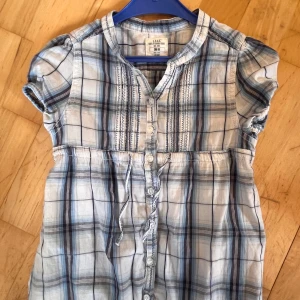 Blårutig kort klänning från L.O.G.G. - Söt kort klänning från L.O.G.G. i blårutigt mönster med knappar framtill och dekorativa sömmar vid bröstet. Klänningen har korta puffärmar och ett knytband i midjan. Perfekt för dig som gillar en chill och avslappnad stil.