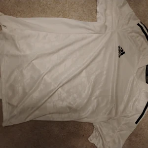 Vit Adidas fotbollströja med svarta detaljer - Säljer en vit fotbollströja från Adidas med svarta ränder på axlarna och Adidas-logga på bröstet. Tröjan är tillverkad i ett lätt och glansigt material som andas, perfekt för träning eller match.