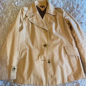 Beige kort trenchcoat från Tommy Hilfiger - Snygg beige trenchcoat från Tommy Hilfiger med klassisk krage, stora knappar och quiltat foder. Jackan har långa ärmar med knappdetaljer och två fickor framtill. Perfekt för dig som gillar stilren och tidlös design. Storlek L men passar mer som en M. 
